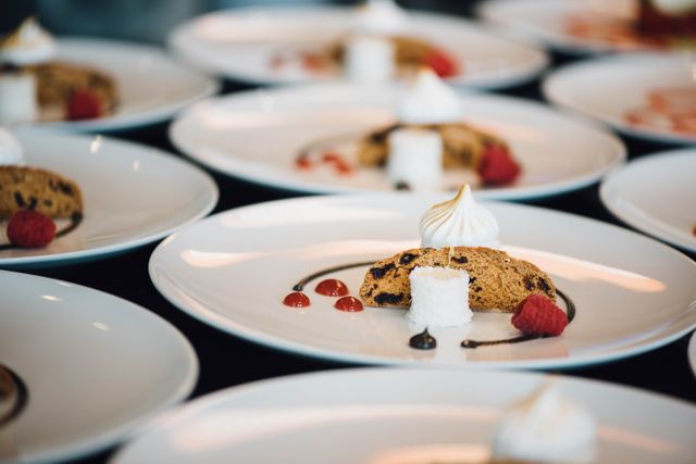 This visually appealing image portrays gourmet dessert plates arranged meticulously for a culinary event. The focus composition highlights intricate food details with artistic presentations involving cookies, dollops of whipped cream, raspberries, and decorative sauces. Perfectly suited for use in culinary magazines, restaurant marketing materials, chef portfolios, or visual narratives related to fine dining and gastronomical arts.