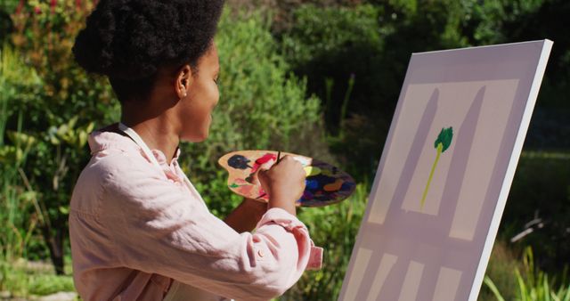 A woman is painting on a canvas in a serene garden setting. She is wearing a pink shirt and an apron, holding a colorful palette. This scene embodies creativity and relaxation, making it ideal for themes of artistic expression, hobbies, leisure, and connecting with nature. Suitable for use in content related to art classes, therapeutic activities, outdoor hobbies, or lifestyle blogs.