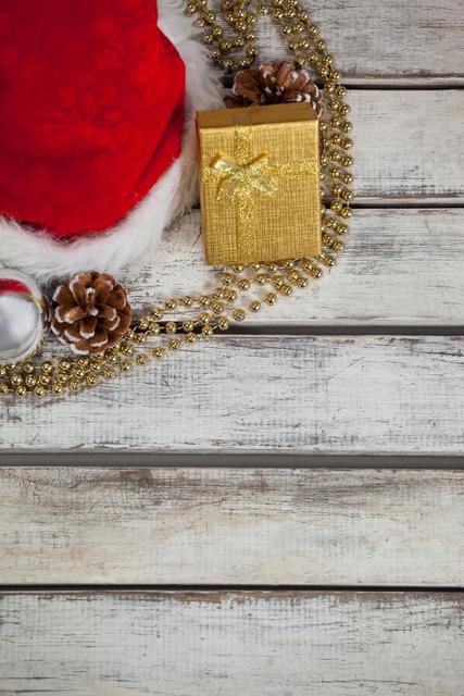 This image of a Santa hat, wrapped gift, golden beads, and pine cones on a rustic wooden table exudes festive charm. Use it for holiday-themed invitations, seasonal social media posts, greeting cards, or Christmas promotional material.