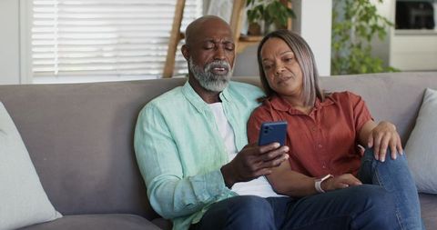 Mature african american couple relaxing and browsing smartphone on living room couch