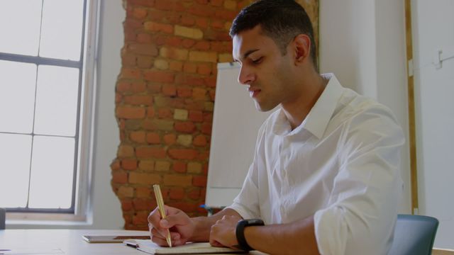 Young businessman focuses intently while writing on diary at his desk in a modern office setting, showing professional dedication. The contrast of a contemporary office and casual attire makes it versatile for usage in materials related to freelance work, modern business cultures, and creative professional environments.