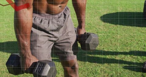 Shirtless athlete holding hex dumbbells during outdoor strength training on grassy field