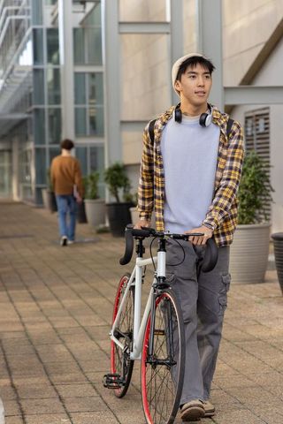 Young asian man pushing road bike on urban campus walkway wearing beanie and headphones