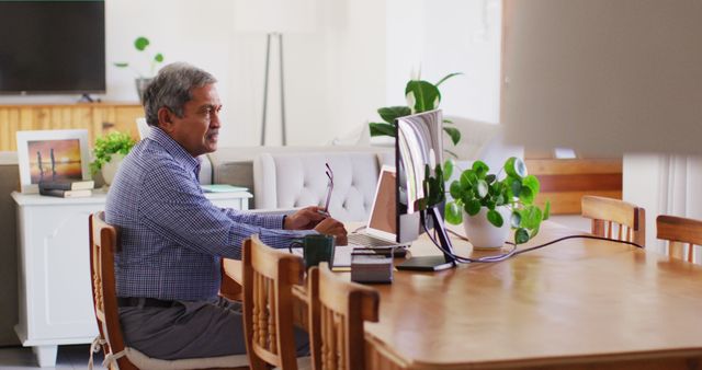 Senior man efficiently working on a laptop at a spacious wooden table in a contemporary home office. The scene features a clean and modern interior with indoor plants and large windows, creating a peaceful and focused environment. Perfect for illustrating concepts like remote work, home office setup, seniors embracing technology, or productive workspace designs.