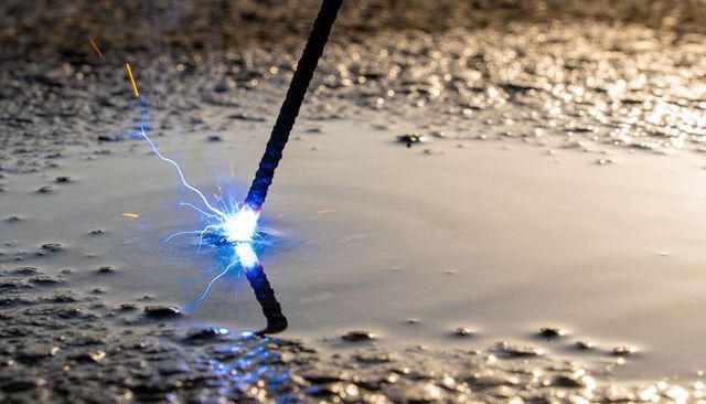 Sparking rod emitting blue-white electrical arc reflecting on muddy puddle surface