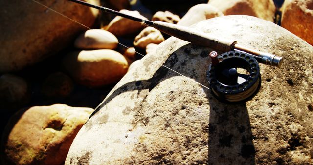 Fishing rod and reel are resting on large sunlit rocks, creating a peaceful outdoor scene. Ideal for use in articles, advertisements, and content related to fishing, outdoor activities, and sport hobbies.