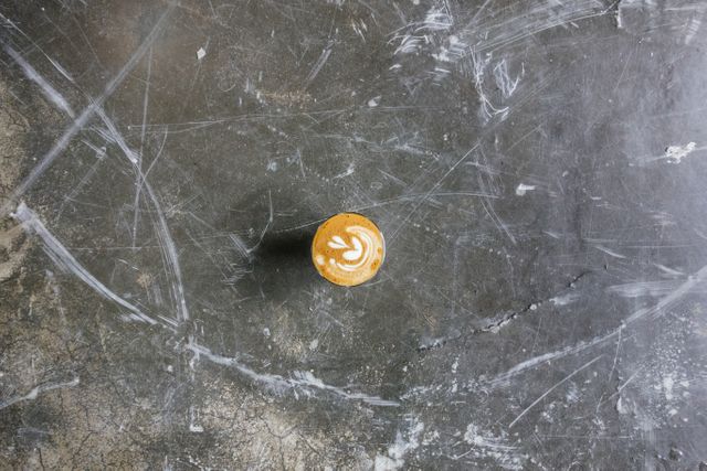 This top down view of a cappuccino on a rustic and distressed concrete surface captures the artistic detail of the latte art in the foam. Ideal for use in coffee shop promotions, lifestyle blogs, food and beverage advertisements, or any content related to coffee culture and morning routines.