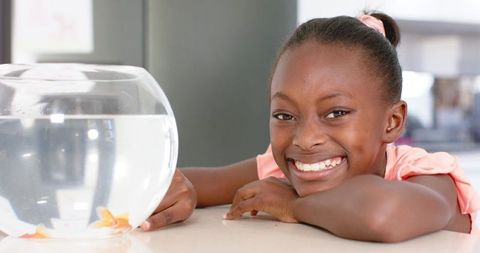 Joyful african american girl smiling while watching goldfish bowl on kitchen counter