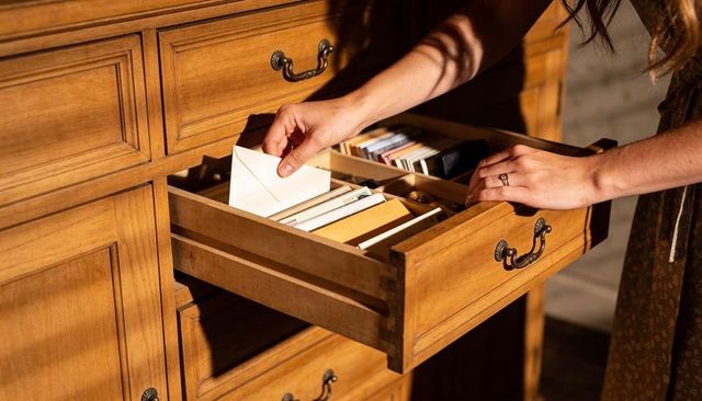 Woman reaching into wooden drawer pulling white envelope sunlit home organization detail