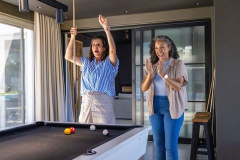 Two diverse women celebrating pool win in modern home gameroom, cheering and clapping