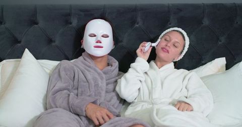 Multicultural friends relaxing in bed during at-home spa night with led mask and facial device