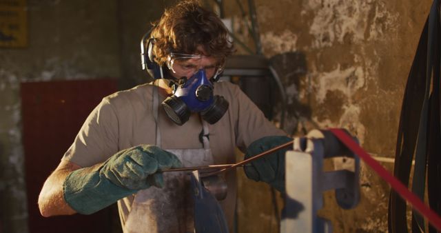 Caucasian male blacksmith wearing breathing mask and ear guards, forging metal tool in workshop. small business and craftsmanship at a blacksmiths forge.
