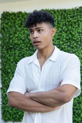 Young man standing with arms crossed wearing white textured shirt against green hedge