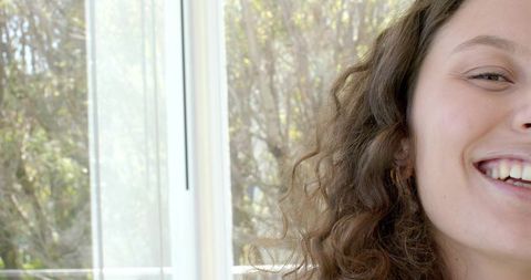 Smiling woman with curly hair by sunlit window showing hoop earring and garden view