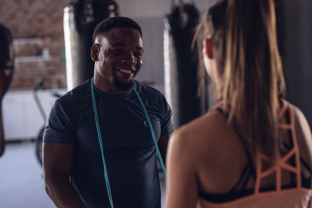 This image shows a smiling trainer with a skipping rope around his neck talking with a young boxer in a gym. Ideal for use in fitness and health promotions, gym advertisements, sports training programs, and motivational content.