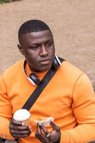 Contemplative african american man sitting wearing orange sweater holding coffee and muffin