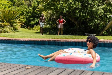 African friends enjoying backyard pool, woman relaxing on pink donut float in sunshine
