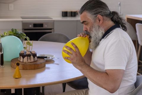 Mature bearded man inflating yellow balloon for intimate home birthday celebration with chocolate dr