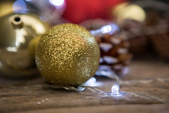 Golden Christmas bauble resting on a wooden plank, surrounded by festive lights and a pinecone. Ideal for holiday greeting cards, festive advertisements, seasonal blog posts, and Christmas-themed decorations.
