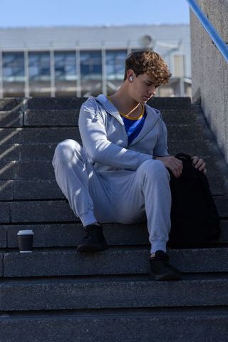 Teen male in gray tracksuit sitting on campus steps listening to earbuds