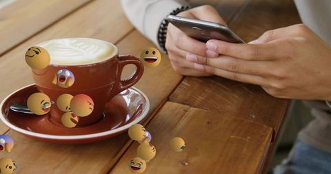 Checking Phone over Latte Art Cup with Floating Emoji Stickers on Rustic Wooden Cafe Table
