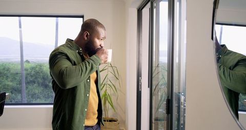 African American man sipping coffee while looking out glass doors in modern home