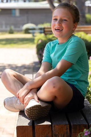 Smiling young girl sitting on bench in sunny park wearing teal shirt and white sneakers