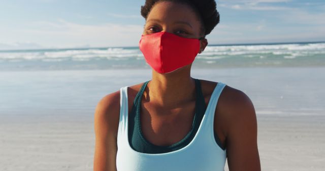 Woman standing on beach wearing red face mask, indicating awareness of health and safety during outdoor activities. Useful for emphasizing health precautions or beach lifestyle during pandemic times, outdoor fitness activities, or promoting beach vacations with safety measures.