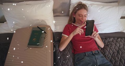 Young woman reclining on hotel bed scrolling smartphone next to suitcase and passport
