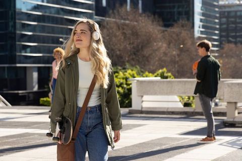 Young woman strolling with skateboard and wireless headphones in modern urban plaza