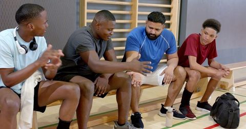 Diverse male teammates discussing strategy and planning play on wooden bench in gym