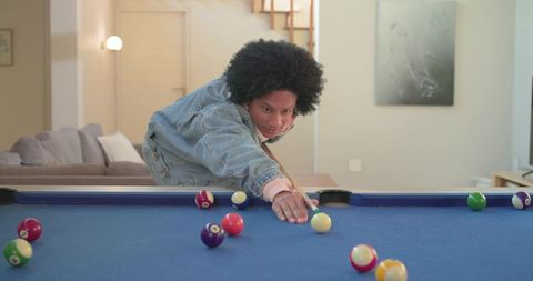 African american man in denim jacket aiming pool cue on blue-felt pool table in modern living room