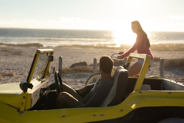 Caucasian woman riding bicycle looking at man in beach buggy on beach at sunset,. beach stop off on summer holiday road trip.