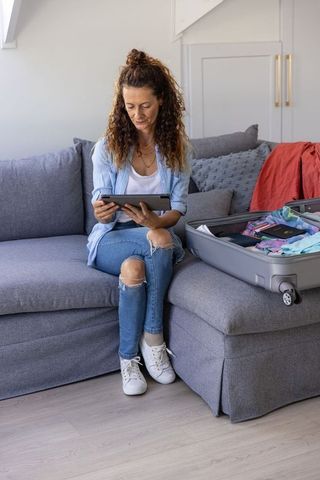 Mature woman packing suitcase with passport while using tablet on gray sofa planning trip