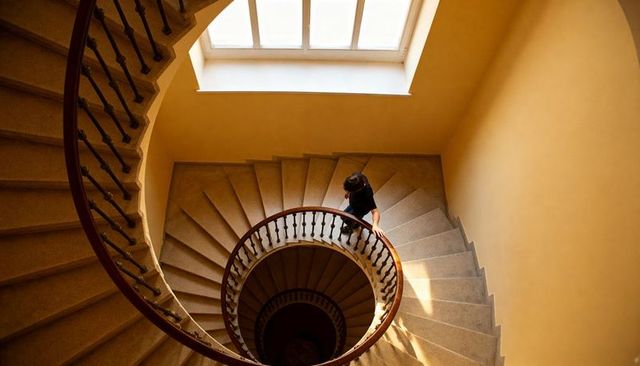 Man descending spiral staircase under skylight in warm-toned stairwell with deep shadows