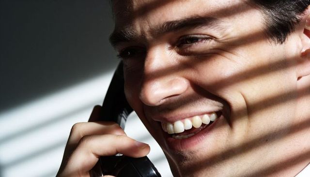 Smiling man talking on corded phone with window blind shadows, closeup portrait