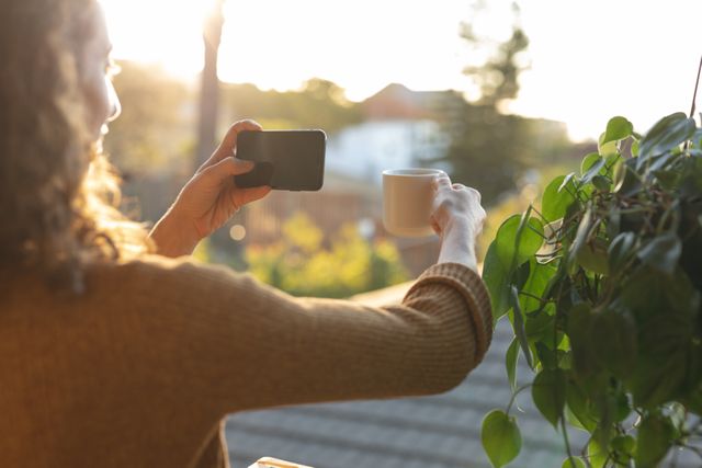 Woman taking photo of coffee cup with smartphone at home during quarantine. Ideal for themes related to home lifestyle, social distancing, relaxation, morning routines, and casual photography.