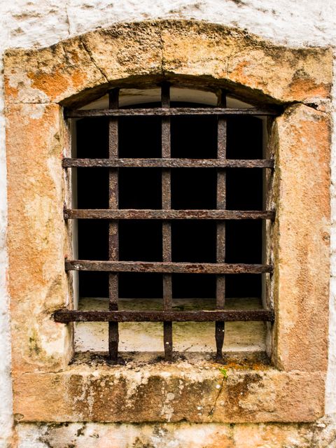 Rusty iron bars covering window with stone frame on old building. Capturing essence of prison, security, and historic architecture. Ideal for articles on history, security equipment, renovating old structures, or exploring themes of imprisonment and isolation.