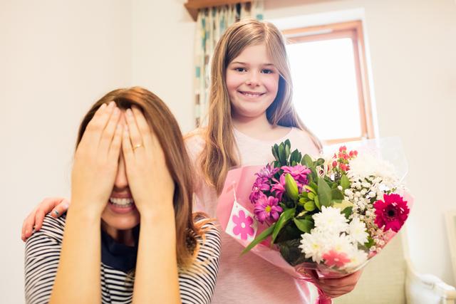 Young girl surprising her mother with a bouquet of flowers in a cozy living room. Perfect for use in family-oriented advertisements, Mother's Day promotions, greeting cards, and articles about family bonding and celebrations.