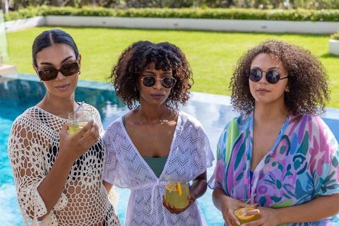 Diverse friends relaxing by pool holding citrus cocktails wearing sunglasses and cover-ups