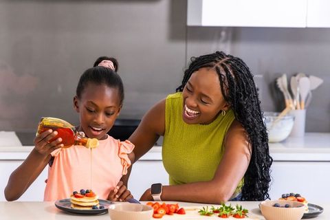 African american mother and daughter pouring syrup on pancakes, laughing during morning breakfast
