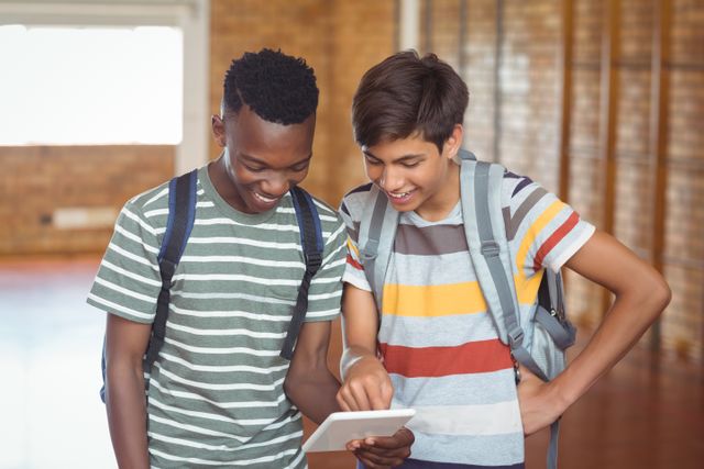 Two schoolboys are standing together in a school hallway, smiling and looking at a digital tablet. Both are wearing casual striped shirts and backpacks. This image can be used for educational materials, technology in education promotions, school brochures, and articles about modern learning and student collaboration.