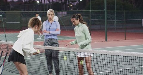 Female tennis coach guiding two players at net during evening practice on outdoor court
