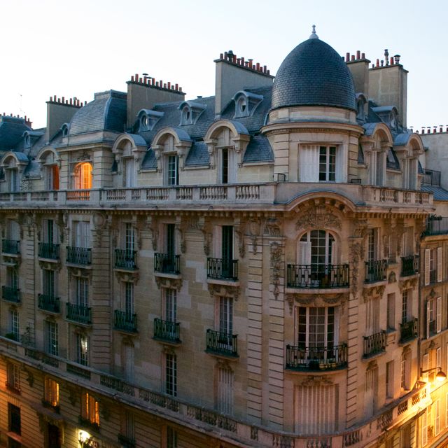 Elegant Parisian apartment building captured at dusk. Detailed with classic French architecture including ornate balconies and a distinctive domed roof. Perfect for travel brochures, architecture blogs, real estate promotions, and articles focusing on urban living in Europe.