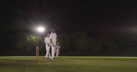 Multicultural male cricketers standing under floodlights preparing to bat on night pitch