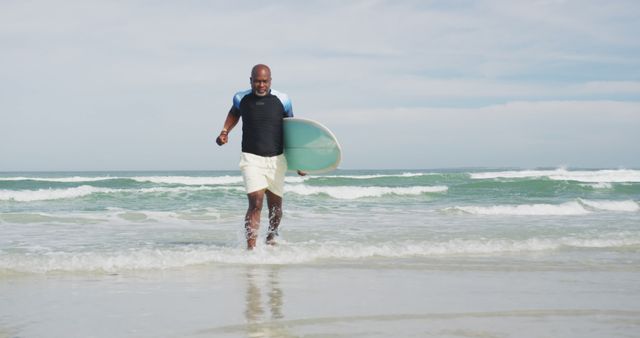 A confident man is emerging from the ocean carrying a surfboard. Useful for topics on adventure, ocean sport, fitness, beach lifestyle, and surfing activities.