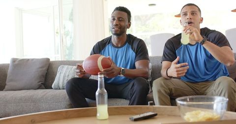 Diverse friends cheering and watching football at home holding ball and drinking beverages