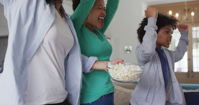 Biracial lesbian couple and daughter watching tv eating popcorn. jumping up in excitement cheering. self isolation quality family time at home together during coronavirus covid 19 pandemic.