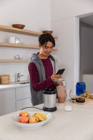 Young asian man blending smoothie in minimalist kitchen while checking smartphone