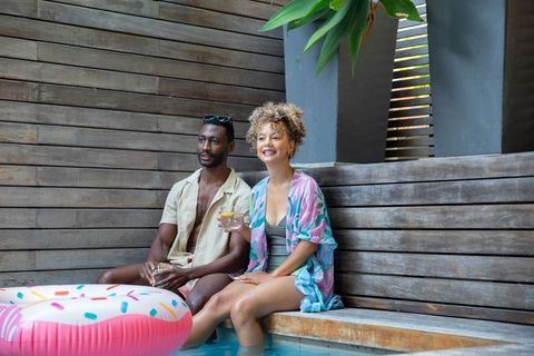 Mixed-race woman and black man relaxing poolside with drinks and colorful donut float